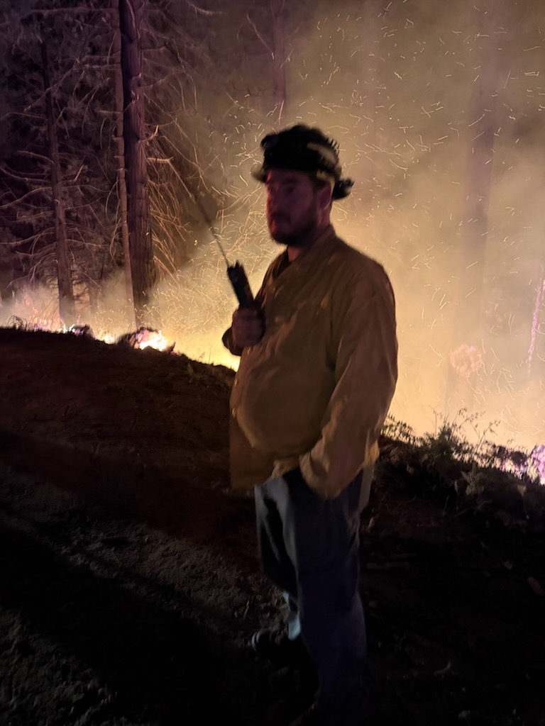 A firefighter standing in front of a wildfire, holding a walkie-talkie, with flames and smoke in the background.