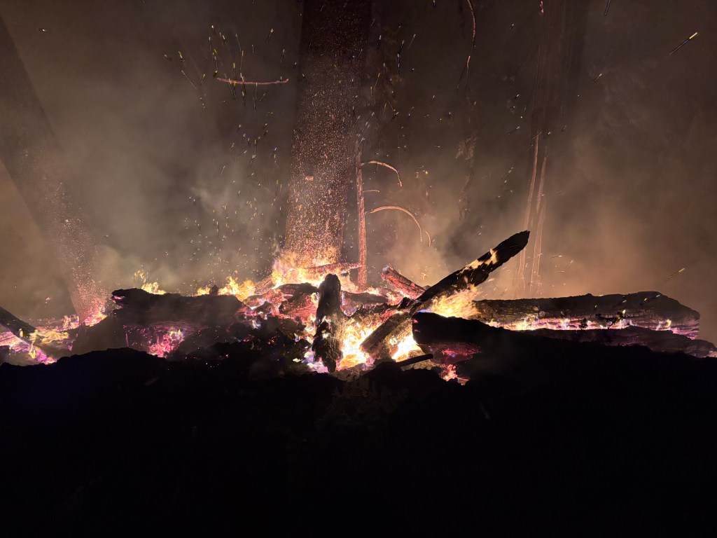 Close-up view of a roaring campfire with glowing embers and smoke filling the air.