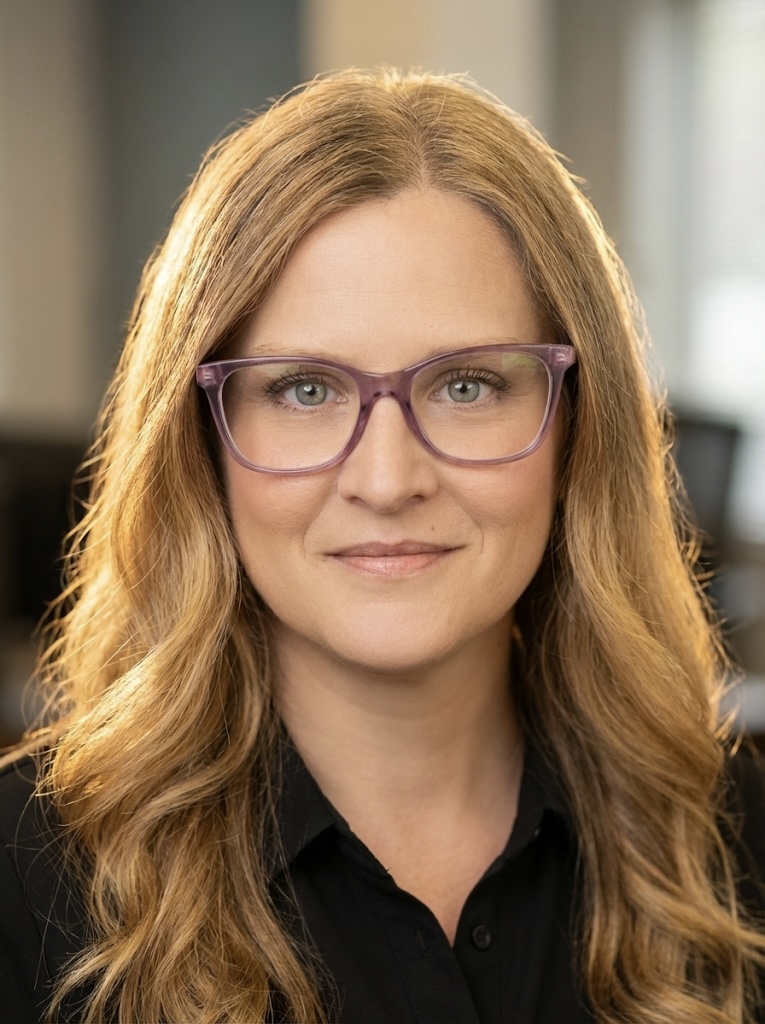 A woman with long, wavy blonde hair and glasses is smiling warmly at the camera. She is wearing a black shirt and is in a softly lit office environment.